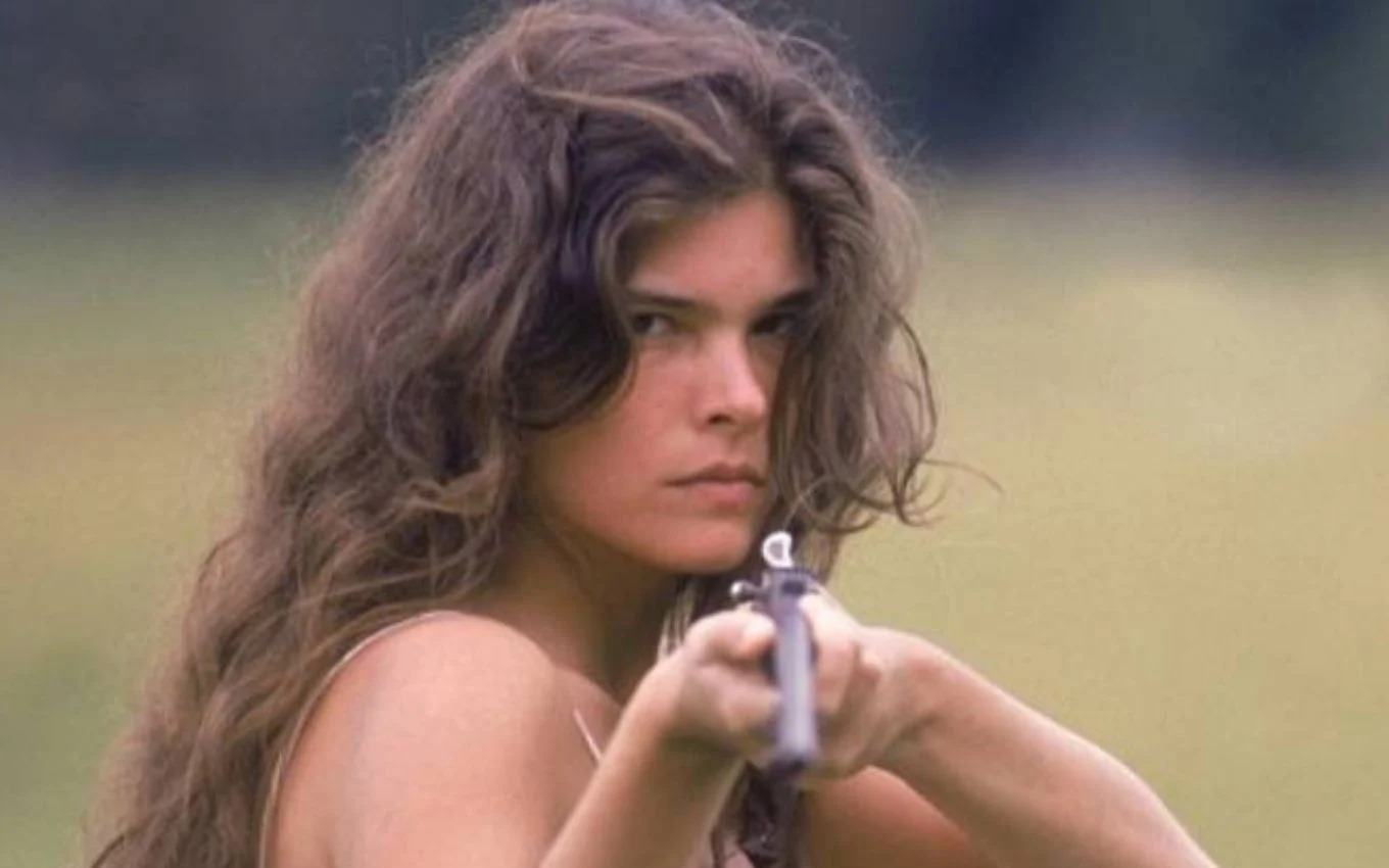 Cristiana Oliveira, como a personagem Juma, segura uma espingarda e mira em um alvo