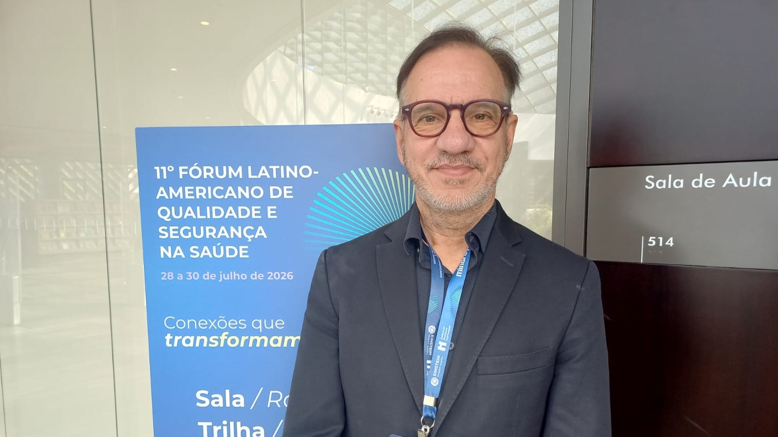 Paulo é um homem baixo, usa óculos e está na frente de um banner