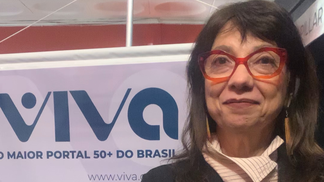 Susanna Lira usa óculos vermelhos e está de pé, na frente do banner do portal Viva