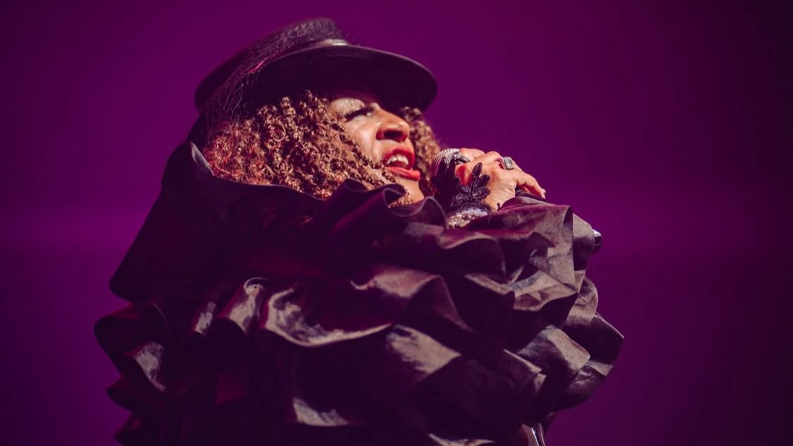 Thea Austin está usando roupa preta, chapéu e está cantando