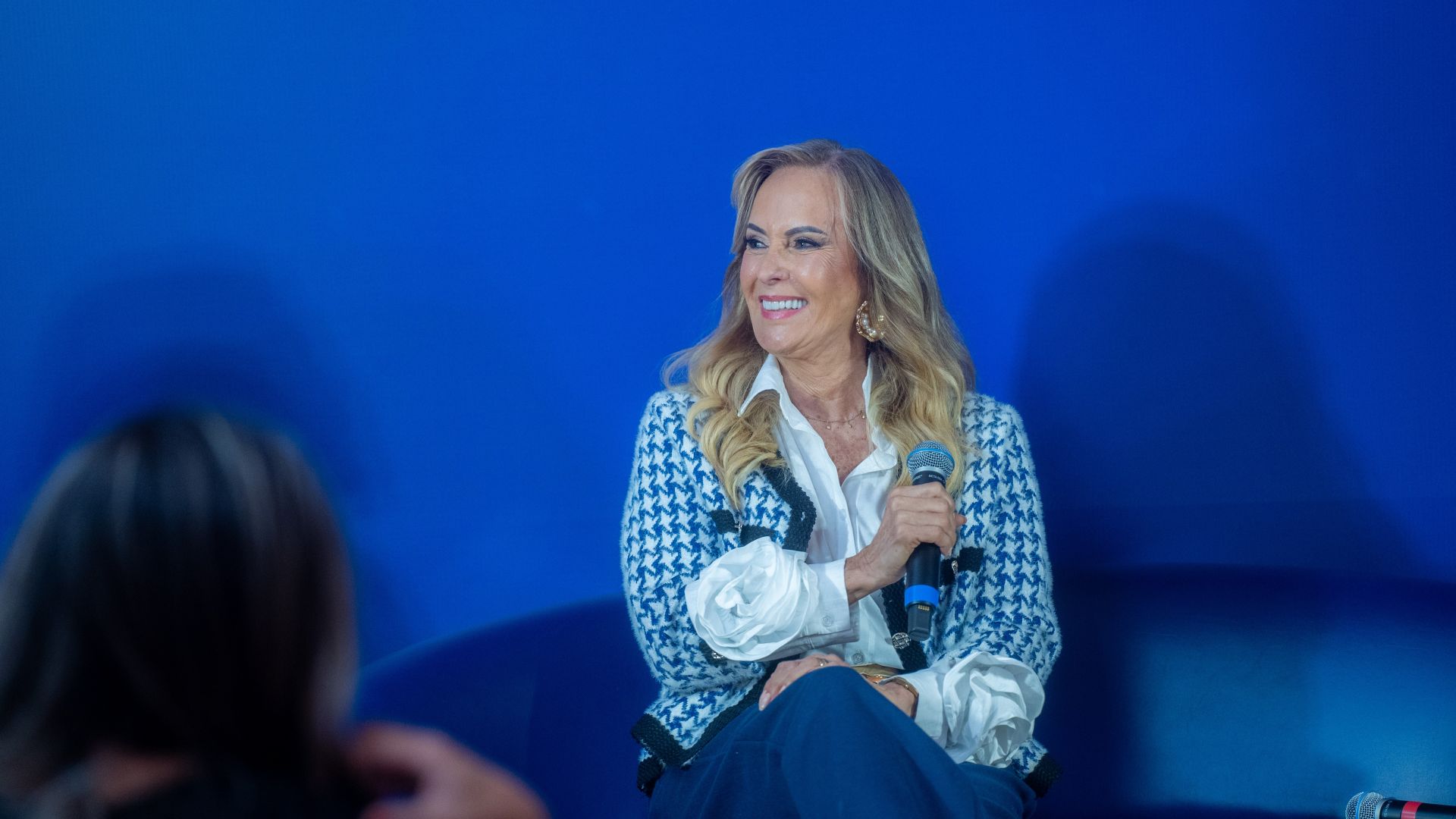 Helô Pinheiro sentada em um sofá segura um microfone sorrindo