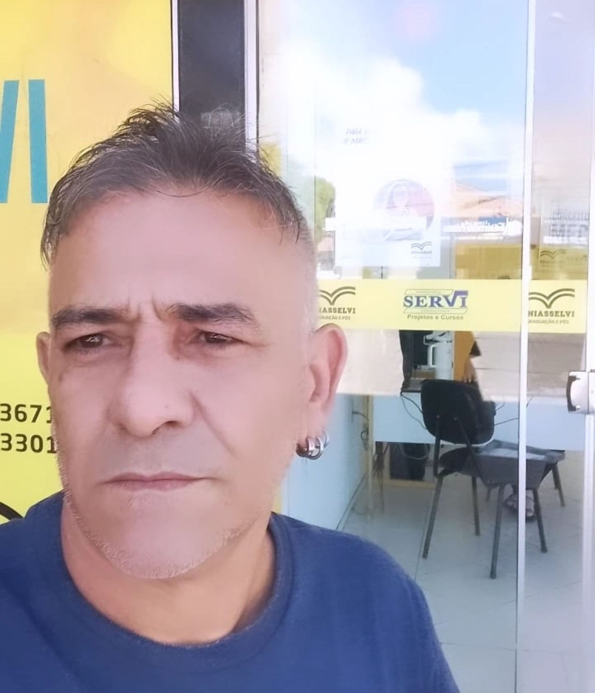 Em área externa, João Amaral está em frente a uma porta de vidro que dá acesso a uma instituição