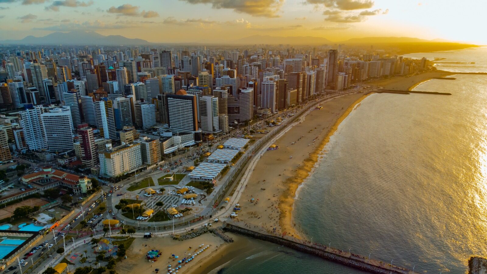Foto aérea da orla de Fortaleza, com a praia e a cidade ao por do sol