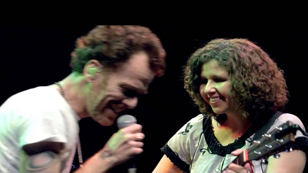 Nando Reis está cantando com microfone enquanto Roberta Campos olha para ele