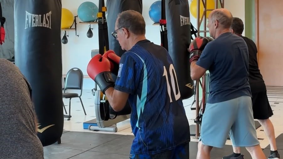 Homens idosos praticam boxe de não contato diante de sacos de pancada na academia.