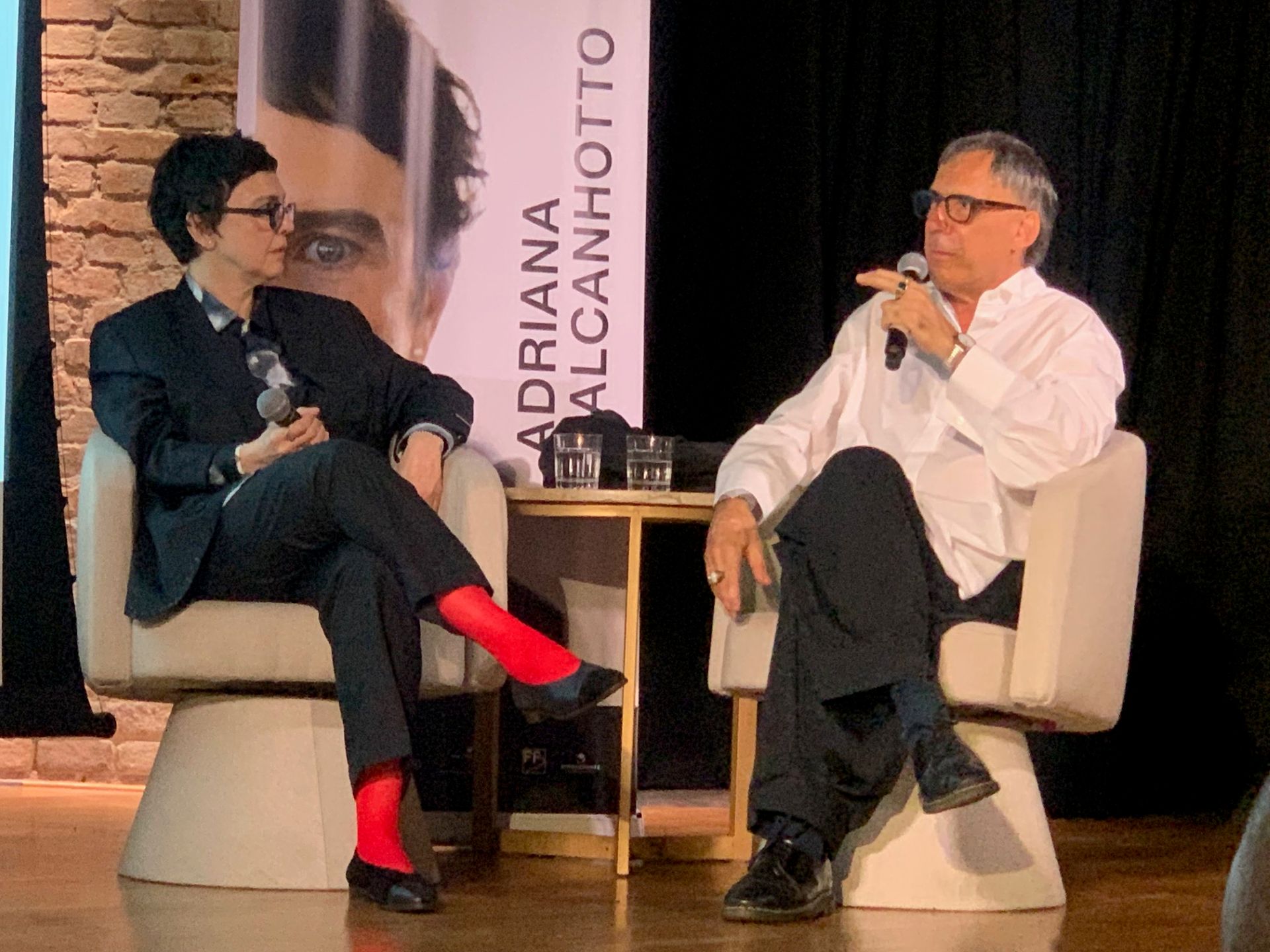 Adriana Calcanhotto e Arnaldo Antunes estão sentados em sofás com microfones na mão, conversando com a imprensa