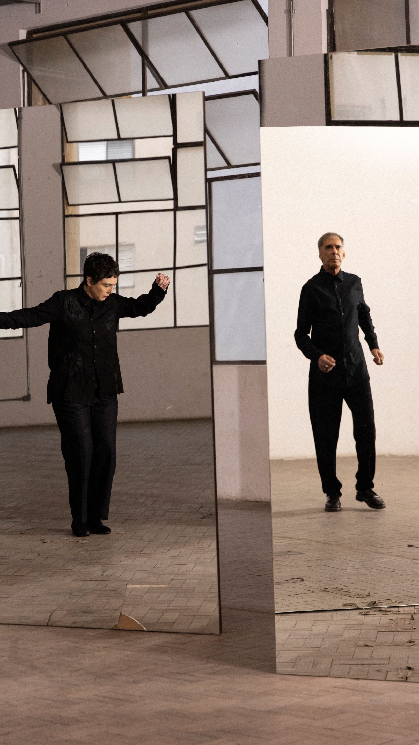Arnaldo Antunes e Adriana Calcanhotto usam roupas pretas e têm suas imagens refletidas em espelhos
