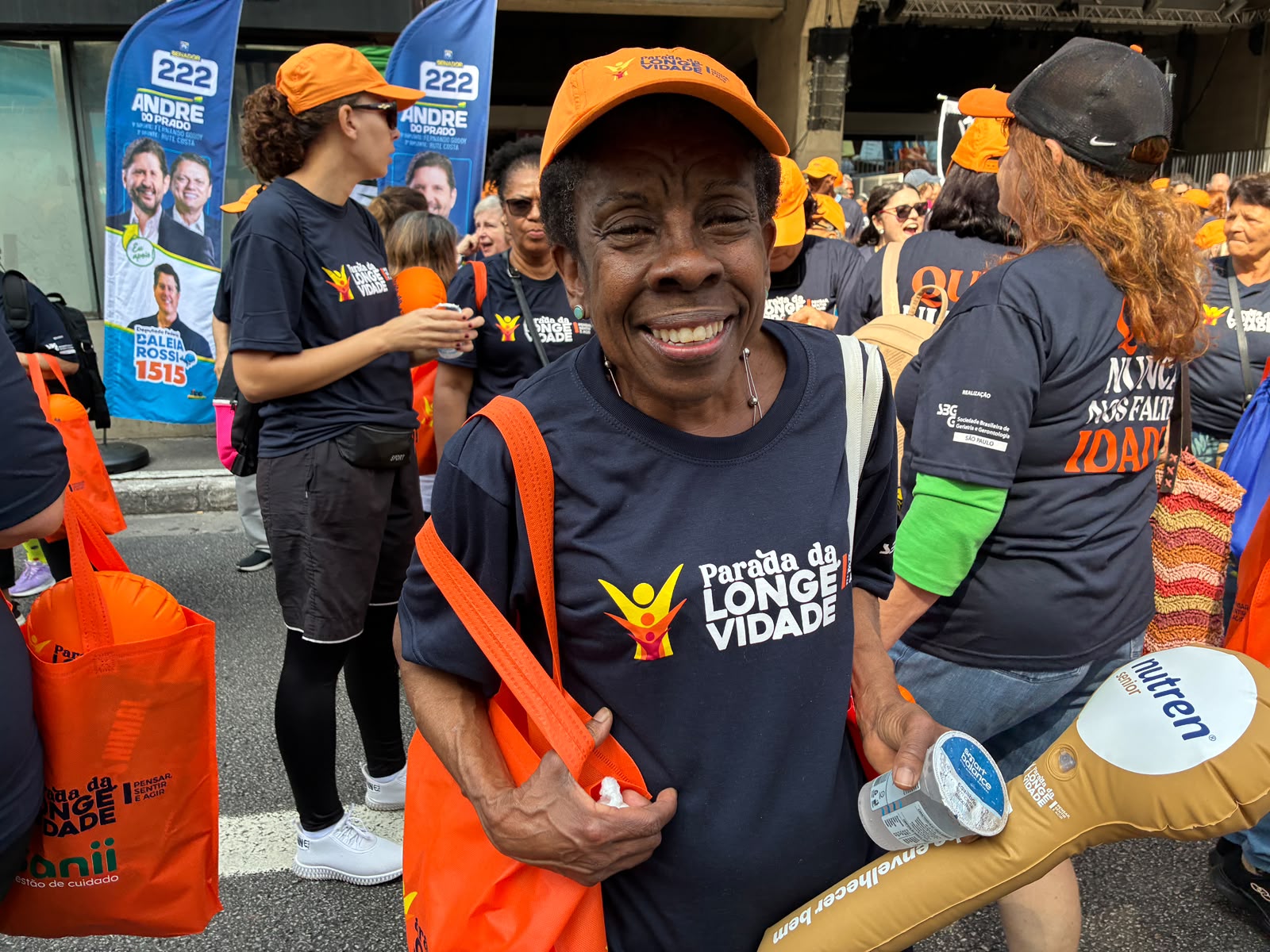 Aposentada Célia de boné laranja sorri na multidão da Parada da Longevidade com banners políticos ao fundo.