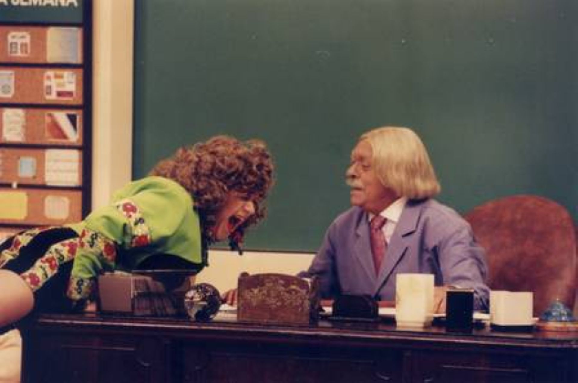 Fafy Siqueira está vestida como a personagem, com roupa verde, em cima da mesa, rindo, sob o olhar de Chico Anysio como o Professor Raimundo