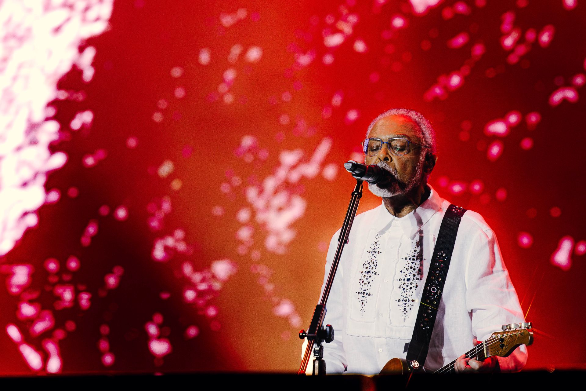 Gilberto Gil usa roupa branco, e o cenário é vermelho