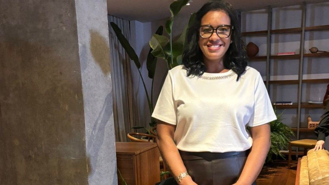 Graziela é uma mulher negra de cabelos escuros e compridos, usa óculos e está de pé em uma sala vestindo uma camisa branca e saia comprida