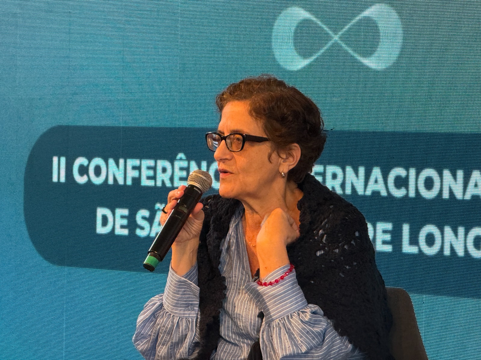 Consultora Maria Cristina Hoffman de óculos e blusa azul fala ao microfone no Fórum da Longevidade.