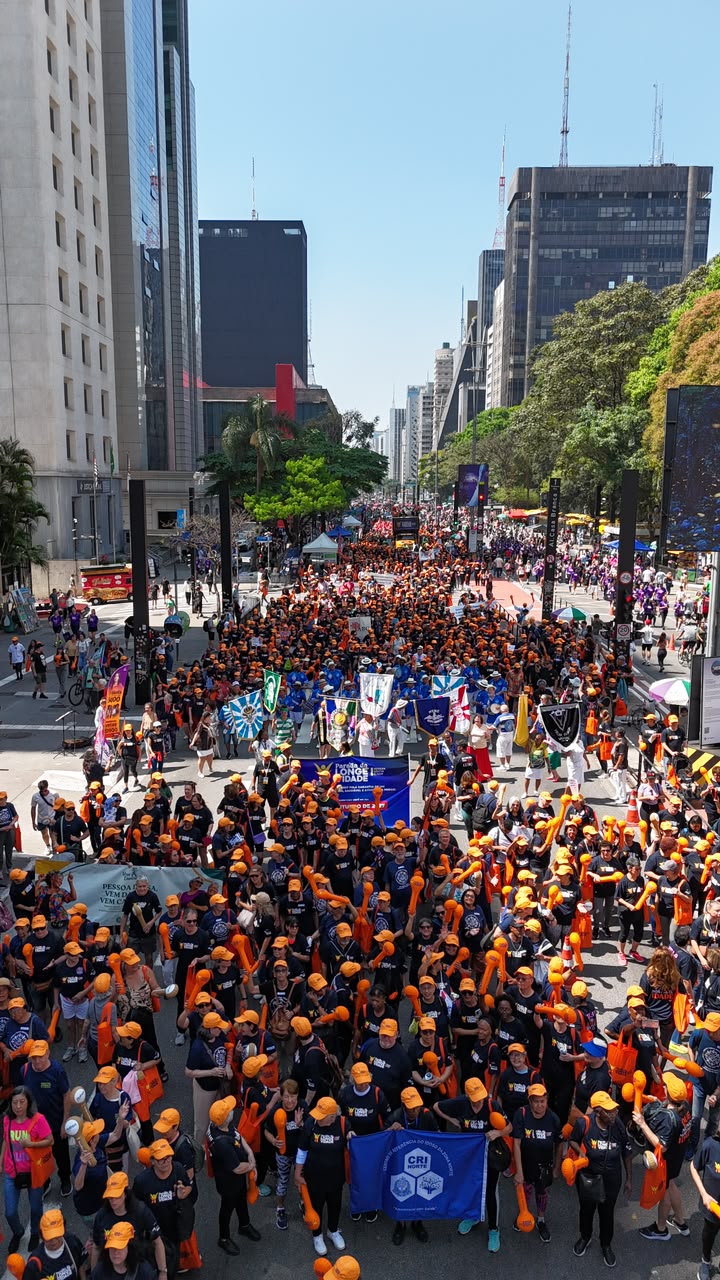 Multidão com camisetas azuis e bonés laranjas caminha na Avenida Paulista durante a Parada da Longevidade.