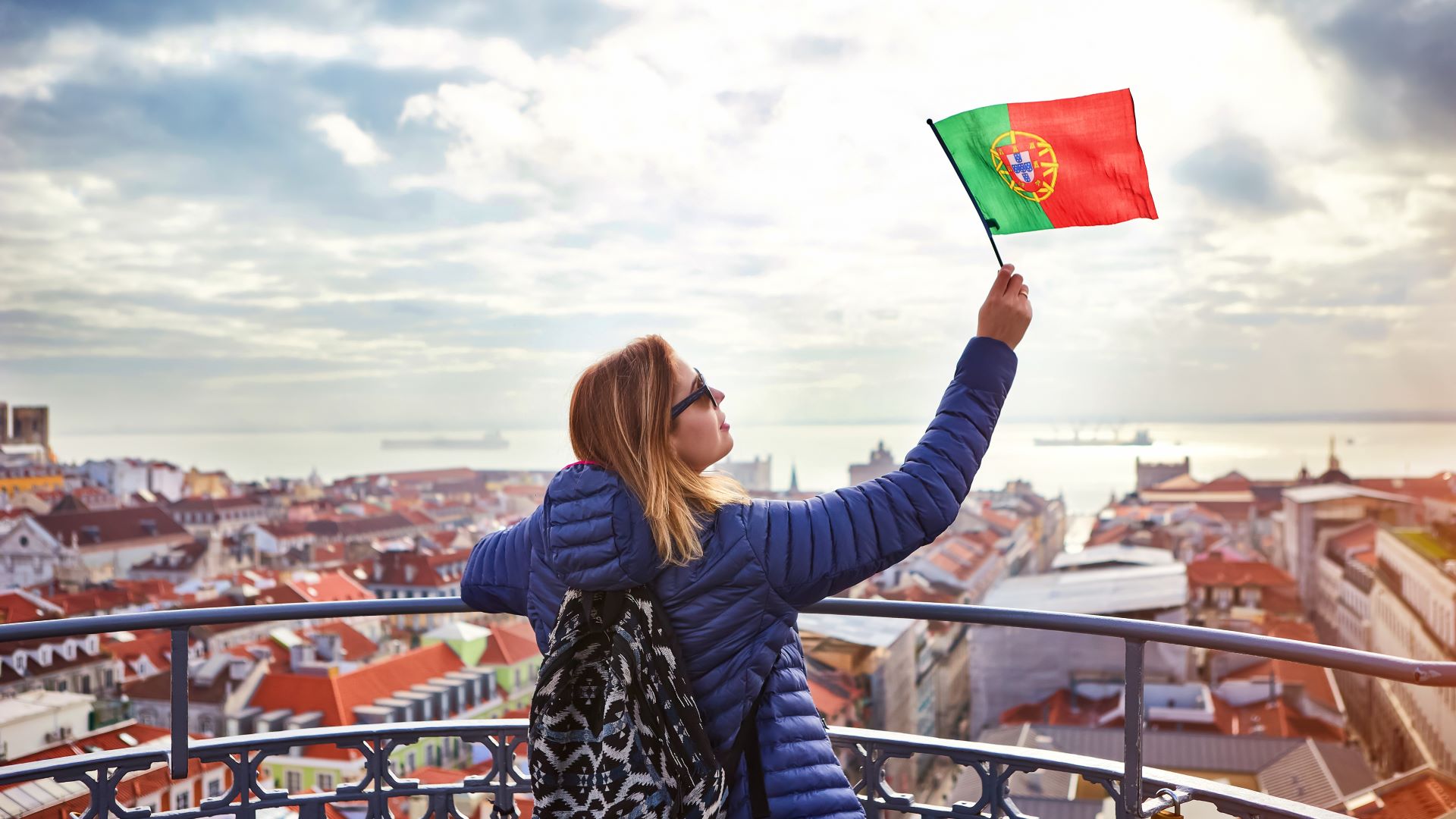 Mulher acena a banbeira de Portugal em um terraço tendo uma vista geral de Lisboa, com o Rio Tejo ao fundo