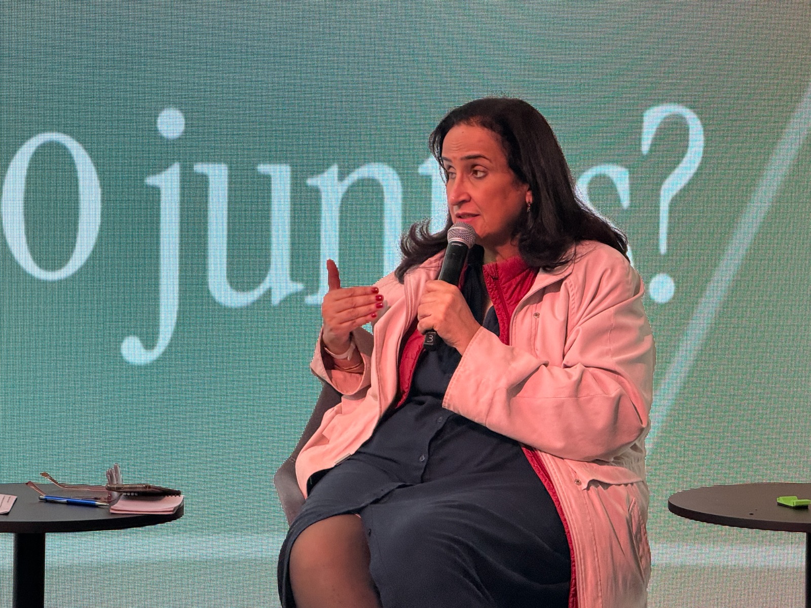 Ativista Regina Helou de casaco rosa fala ao microfone sentada no palco do Fórum da Longevidade.