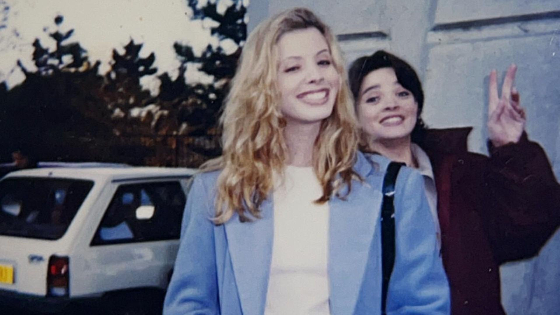 Daniela Valente está com blazer azul sorrindo e Maria Mariana aparece atrás sorrindo também