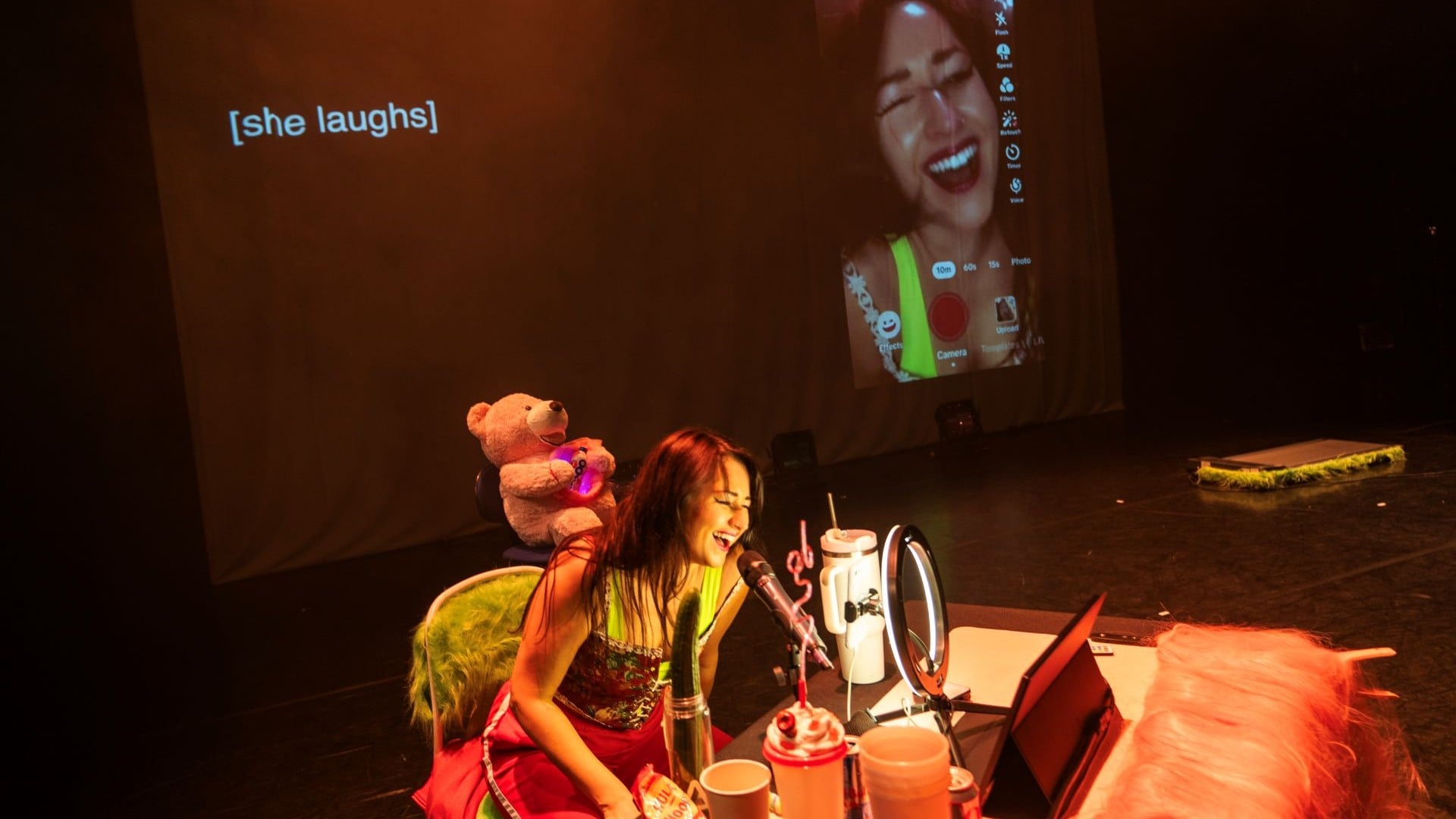 A artista britânica Louise Orwin está cercada de vários objetos em cima do palco e tem um telão atrás dela, em que ela aparece sorrindo