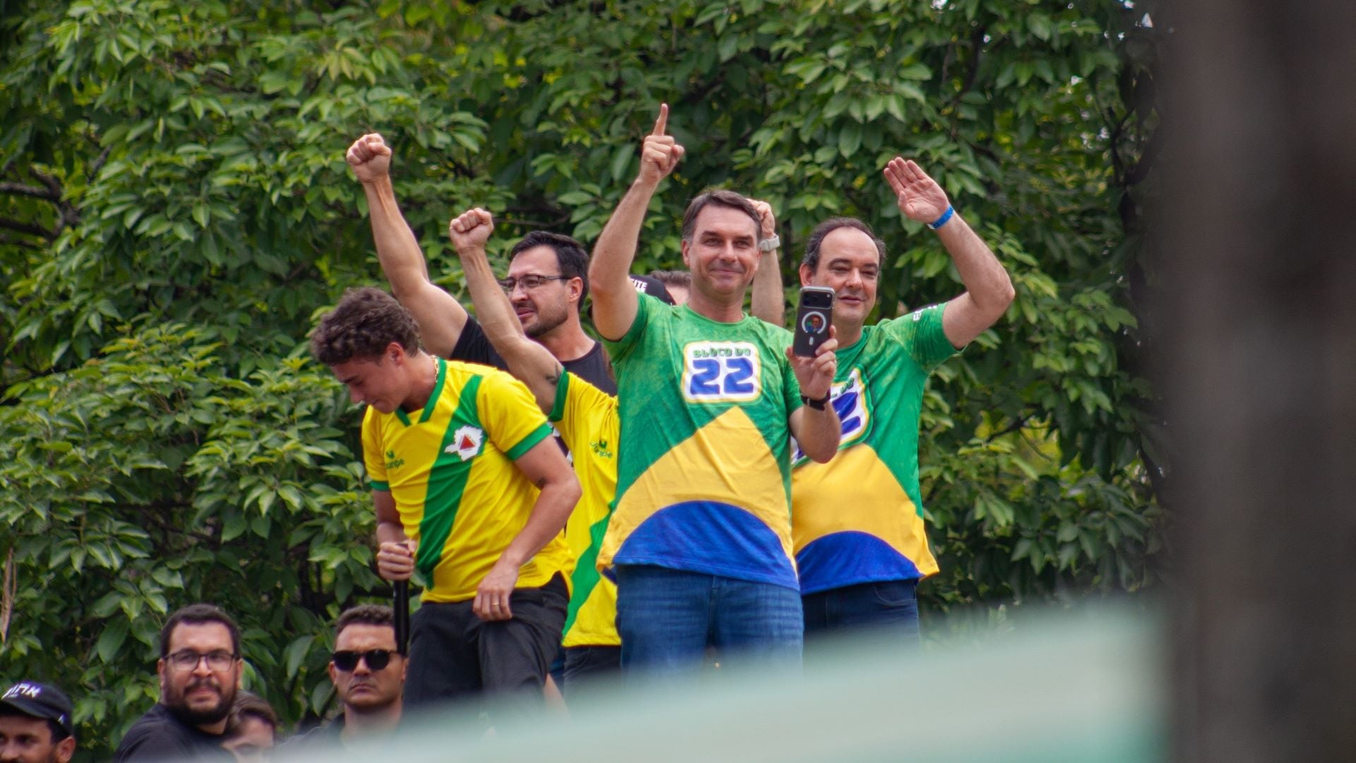 Flávio Bolsonaro e apoiadoras em cima de um carro de som acenam com os braços levantados
