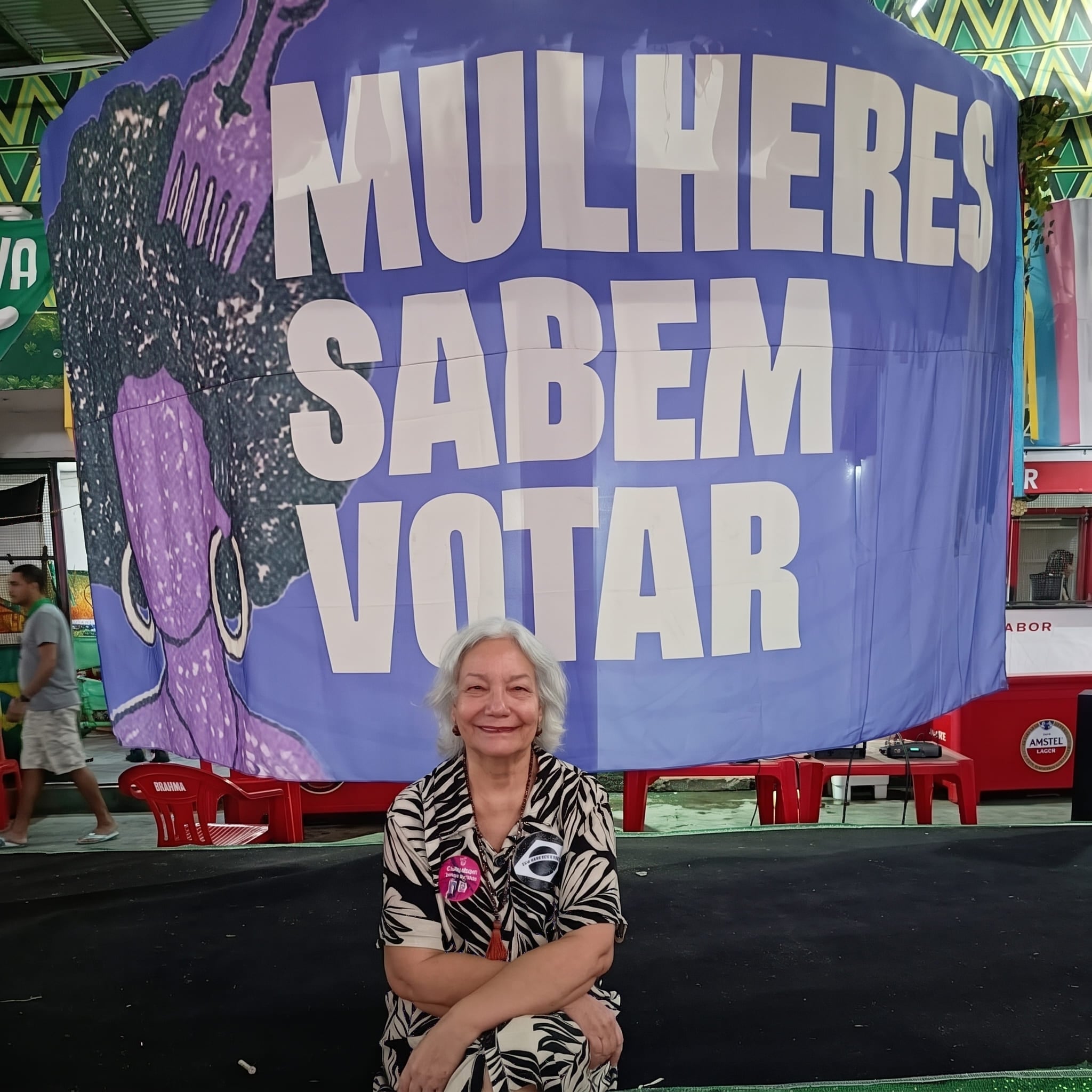 Ativista Genilda Alves sorri diante de grande faixa azul com a frase Mulheres Sabem Votar.