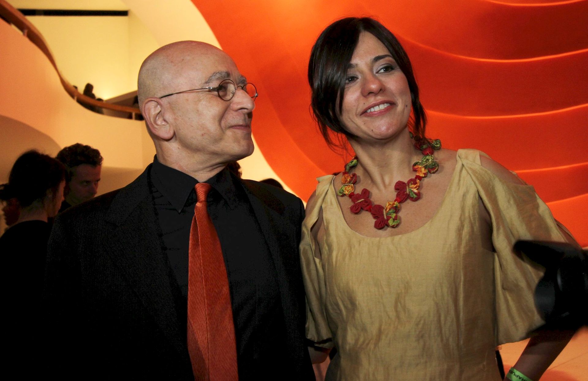 Leon Cakoff usa camisa preta e gravata vermelha e Renata está com vestido amarelo e colocar vermelho