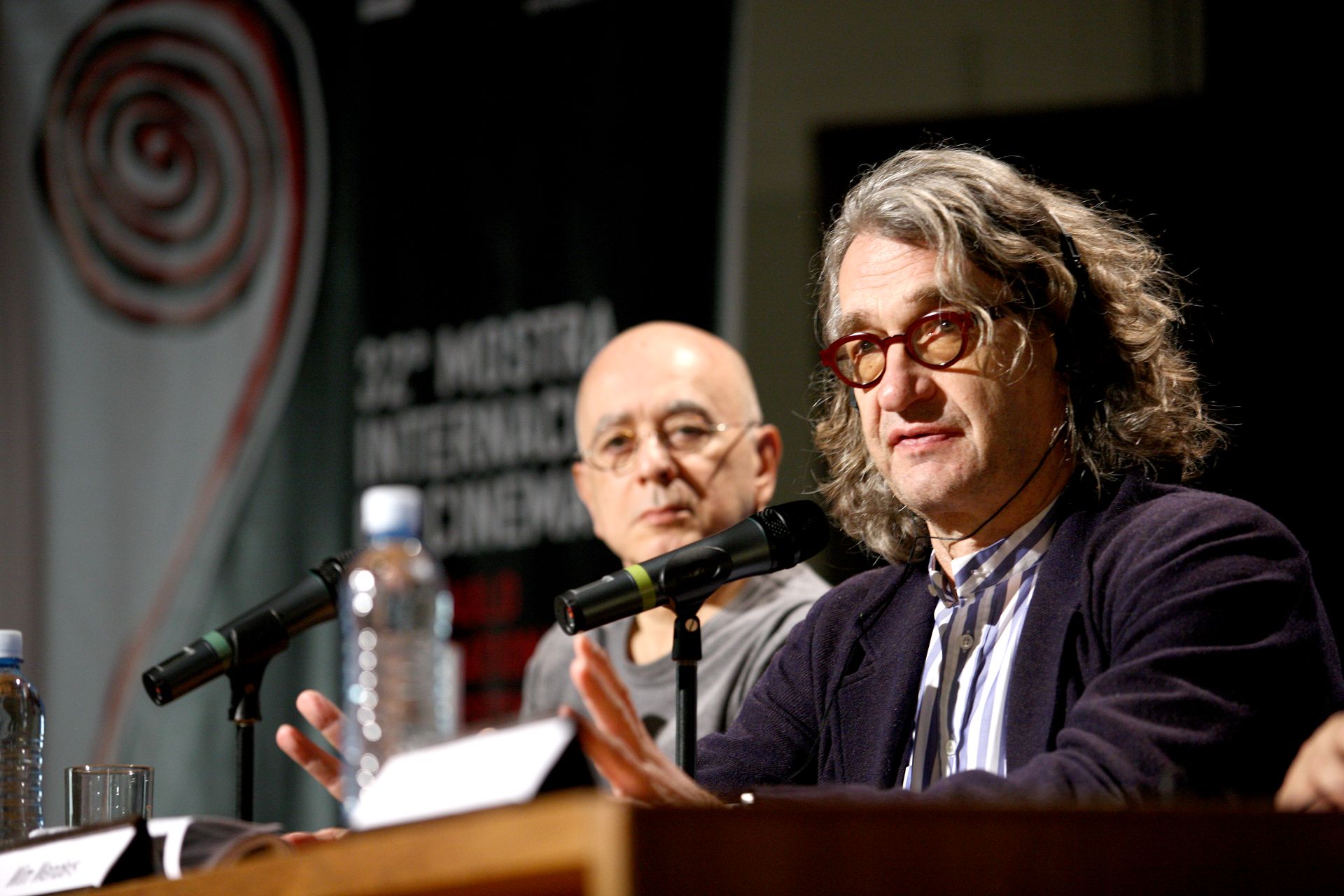 O diretor Wim Wenders está sentado, com um microfone à sua frente e com Leon Cakoff ao fundo
