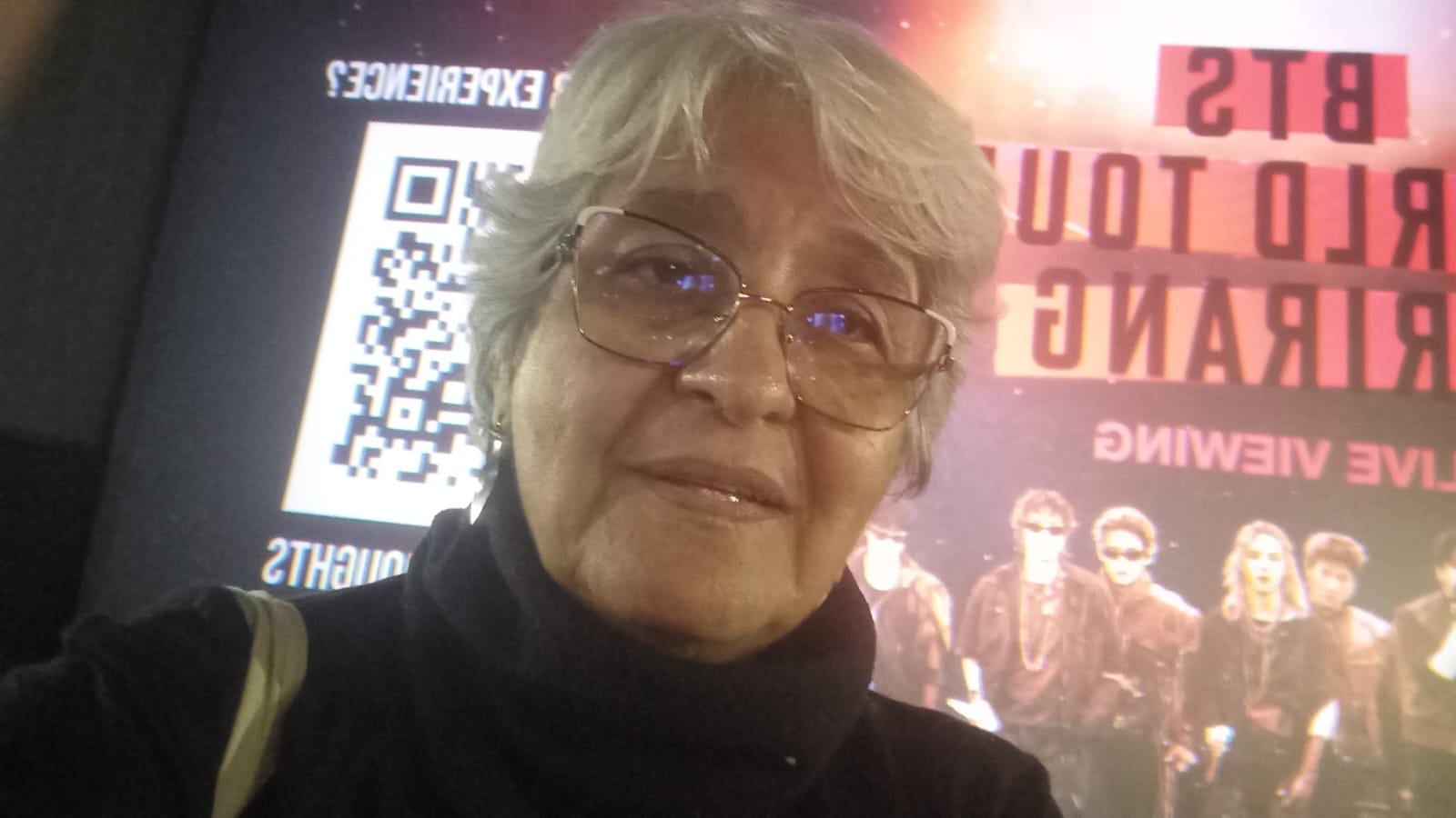 Maria tem cabelos brancos, usa óculos e está na frente de um cartaz do BTS