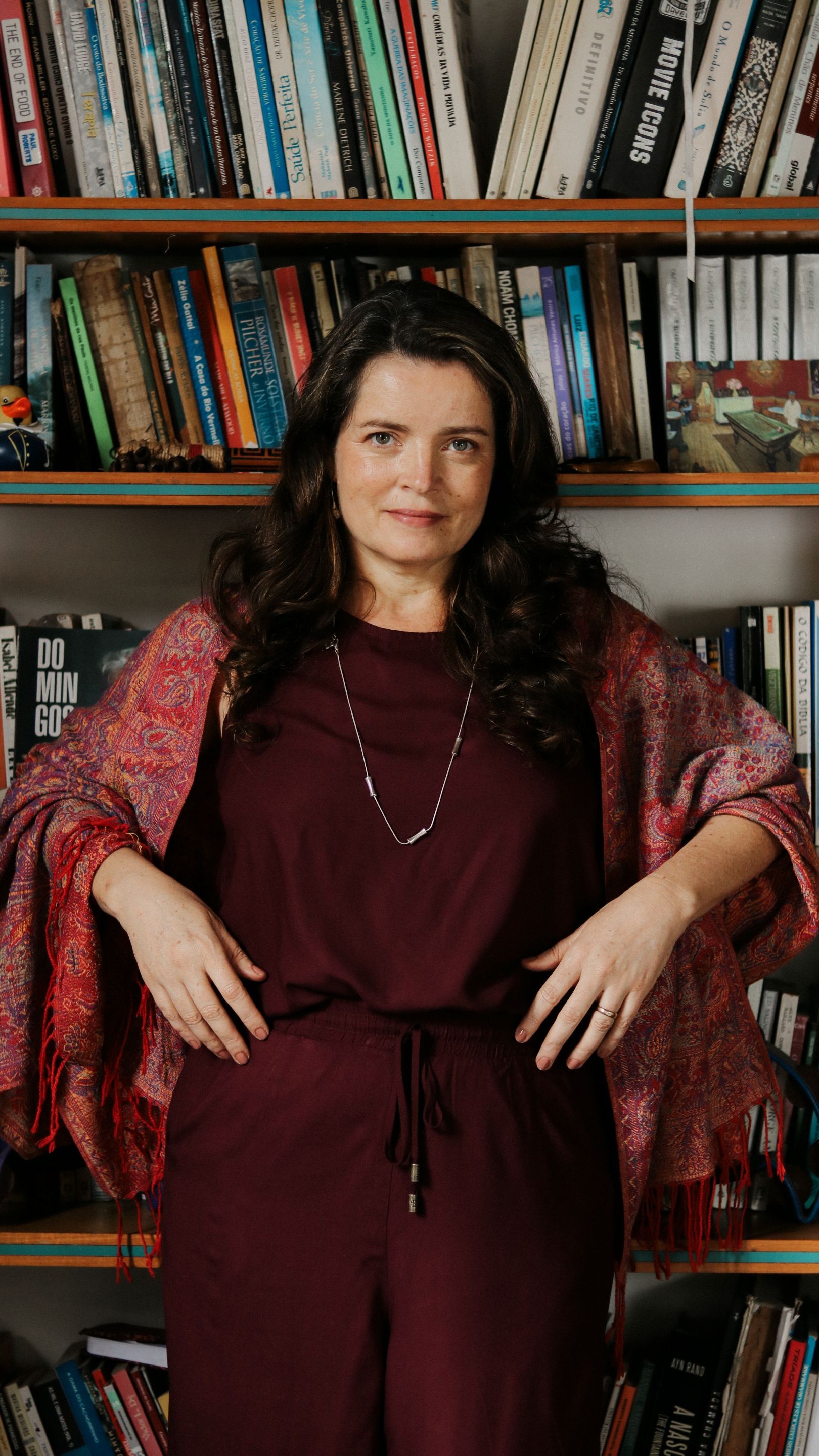 Maria Mariana está de pé, com roupa cor vinho, na frente de uma estante de livros