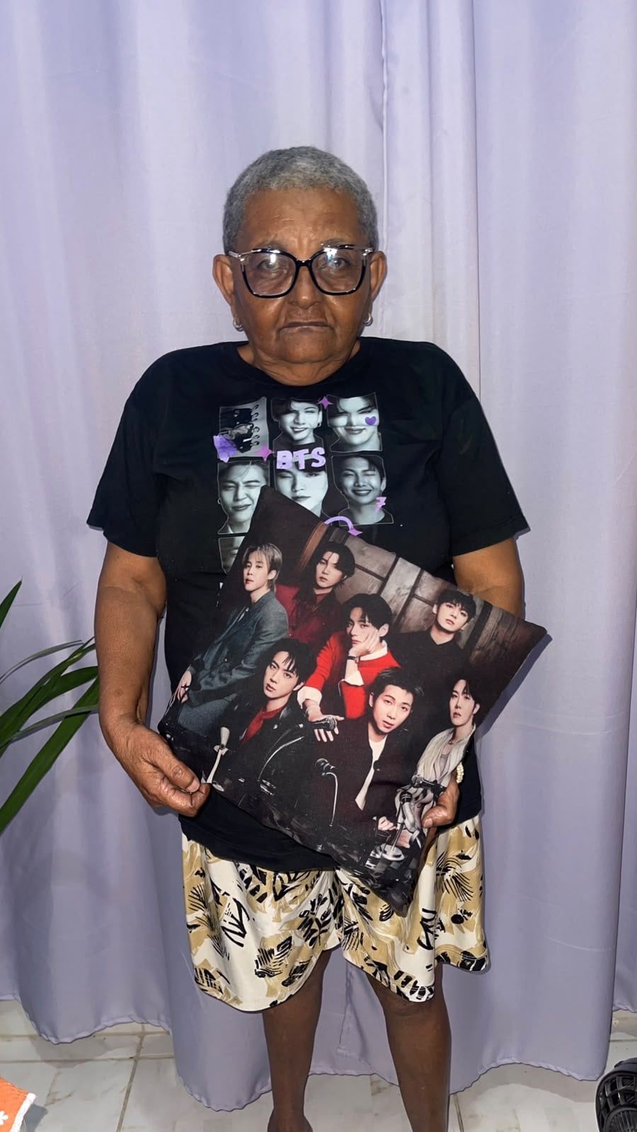 Maria Tânia usa uma camiseta preta estampada com a foto do grupo e segura uma almofada também estampada com o grupo