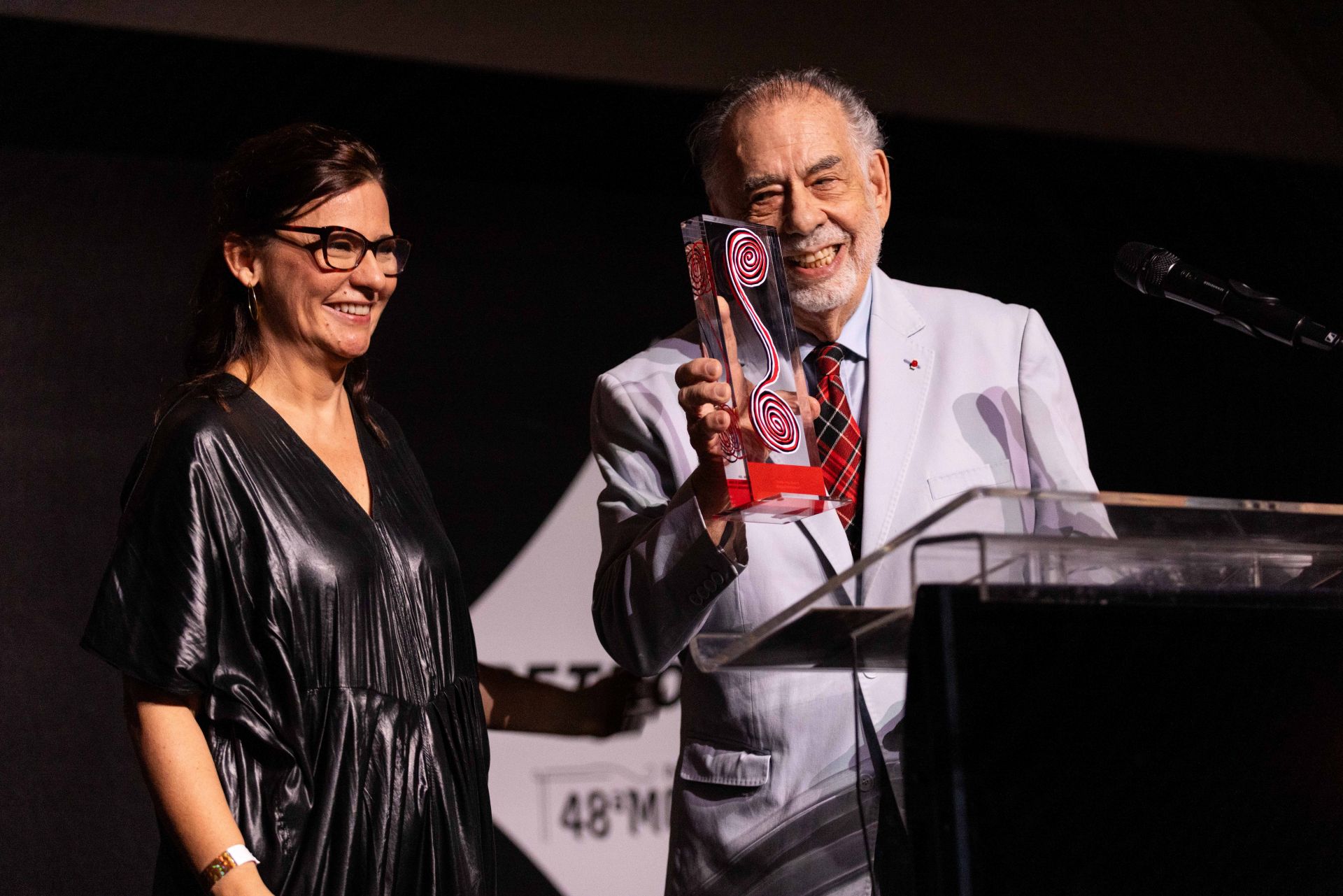 Renata de Almeida está usando um vestido preto e sorrindo, ao lado de Francis Ford Coppola segurando um prêmio