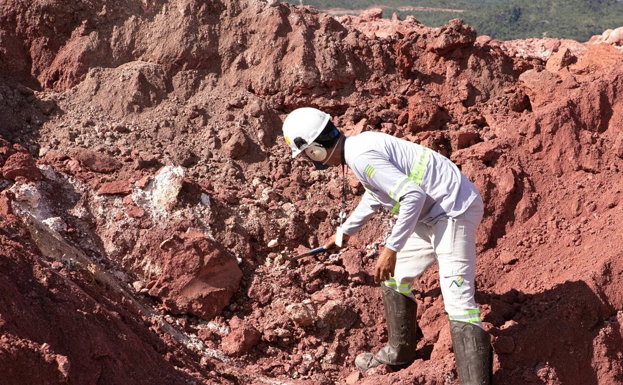 Trabalhador de capacete bate martelo geológico em solo rochoso avermelhado de mineração de terras raras.
