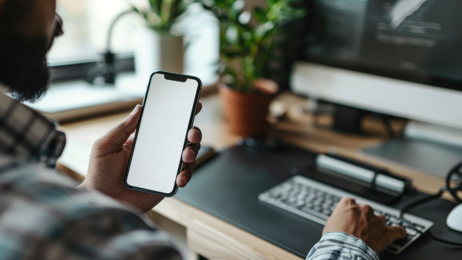 Pessoa digita teclado de um computador com a mão direita e confere o celular segurado pela mão esquerda