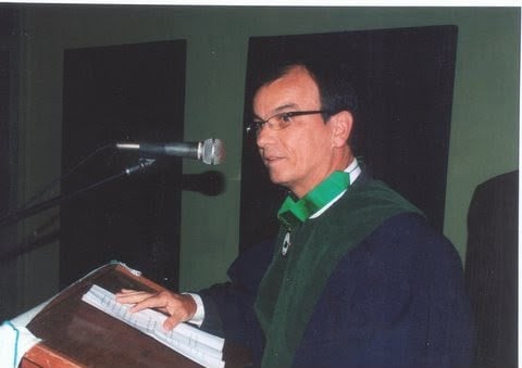 Médico Renato Peixoto Veras discursa ao microfone em púlpito com veste acadêmica.
